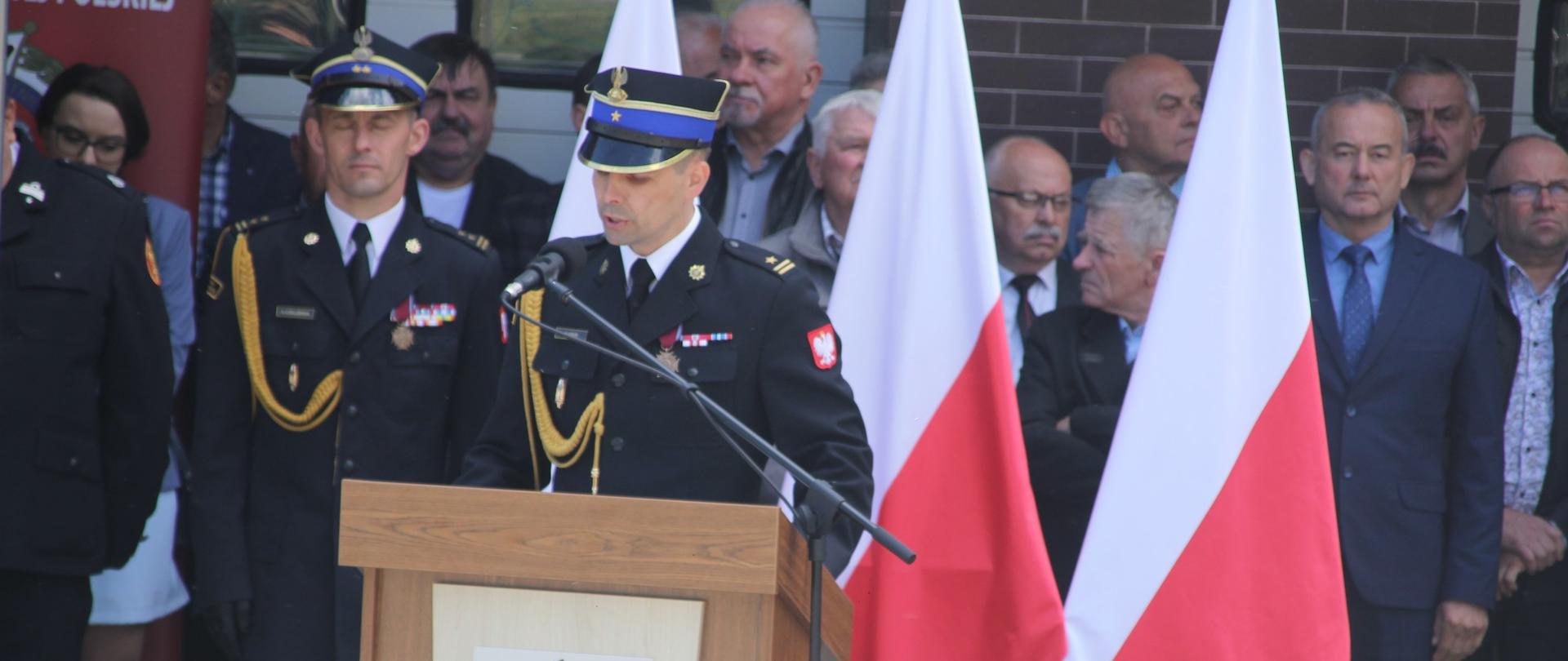 Na zdjęciu widoczny komendant powiatowy Państwowej Straży Pożarnej w Gorlicach przemawiający podczas obchodów powiatowych dnia strażaka w Gorlicach. W tle widoczni zaproszeni goście na uroczystość druhowie OSP i strażacy PSP wszyscy w mundurach galowych i rogatywkach oraz emeryci i renciści pożarnictwa. 