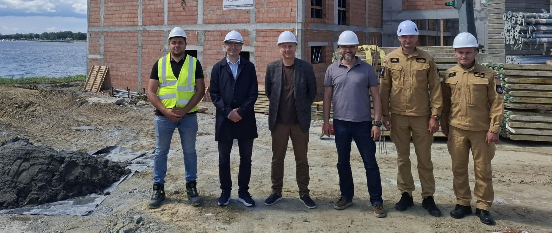 Zdjęcie wykonano na terenie i na tle budowy Podkarpackiego Centrum Ratownictwa i Koordynacji Transgranicznych Działań Ratowniczych w Zakresie Zwalczania Zagrożeń dla Środowiska w Tarnobrzegu w pobliżu Jeziora Tarnobrzeskiego.
Na pierwszym planie znajduje się 6 osób. Od lewej: Konrad Cideł – kierownik budowy, Andrzej Słodki – kierownik Wspólnego Sekretariatu Programu Interreg NEXT Polska – Ukraina 2021-2027, Marcin Kołnierzak – Dyrektor Centrum Projektów Europejskich, Karol Lusar Zastępca Dyrektora Centrum Projektów Europejskich, bryg. Daniel Myszka – komendant miejski PSP w Tarnobrzegu oraz bryg. Witold Nycz – Zastępca Naczelnika Wydziału Logistyki w Komendzie Wojewódzkiej PSP w Rzeszowie. Wszyscy na zdjęciu na głowach mają kaski ochronne, natomiast funkcjonariusze występują w umundurowaniu służbowym w kolorze piaskowym.
