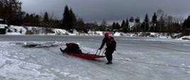 Zdjęcie przedstawia przemyskich strażaków podczas ćwiczenia z zakresu ratownictwa wodno-lodowego , trzech strażaków w skafandrach suchych i kaskach ochronnych w trakcie transportowania strażaka-pozoranta na saniach lodowych