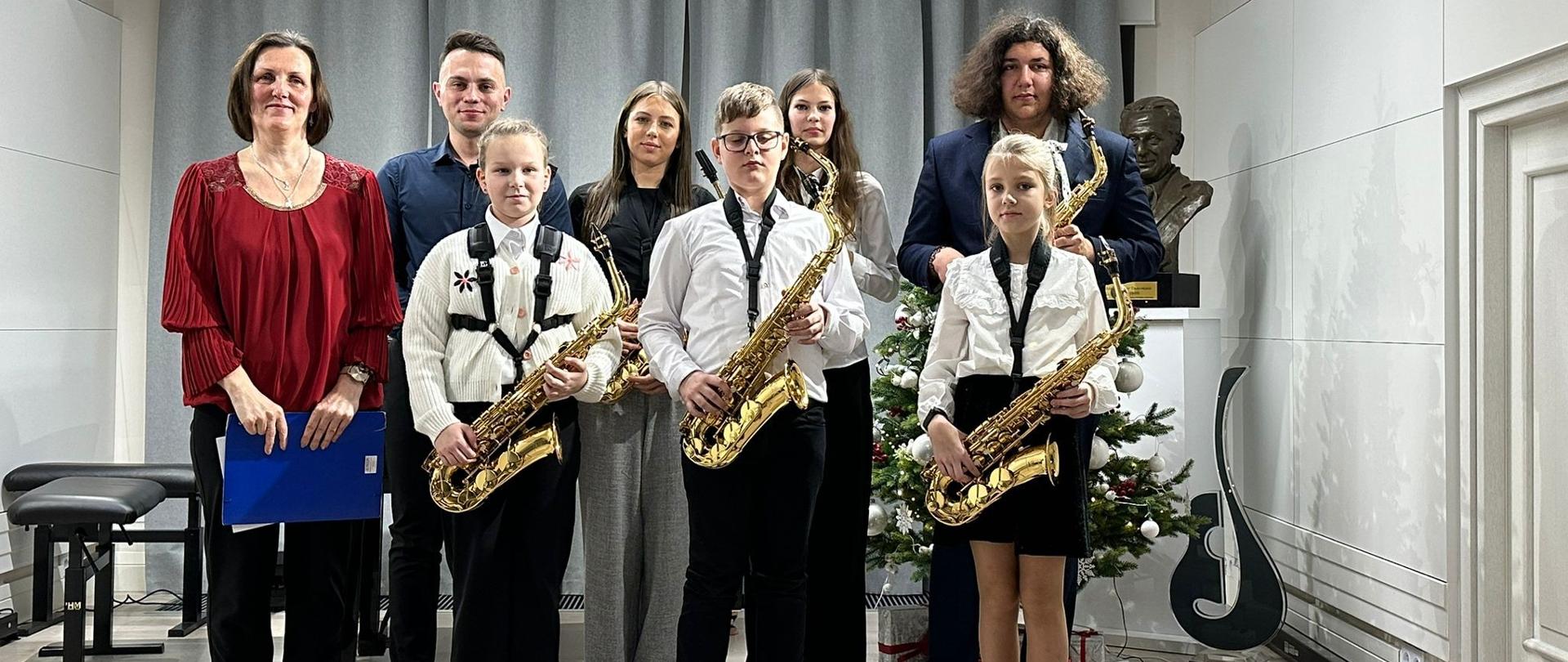 Na zdjęciu widoczna jest grupa uczniów grających (trójka z nich trzyma w rękach saksofony), z nauczycielami. Wszyscy stoją na podium w jasno urządzonym pomieszczeniu koncertowym. Uczniowie ubrani są elegancko: białe koszule, czarne spodnie lub spódnice, a instrumenty zawieszone mają na paskach na szyi. W tle znajduje się ozdobiona choinka oraz popiersie patrona. 