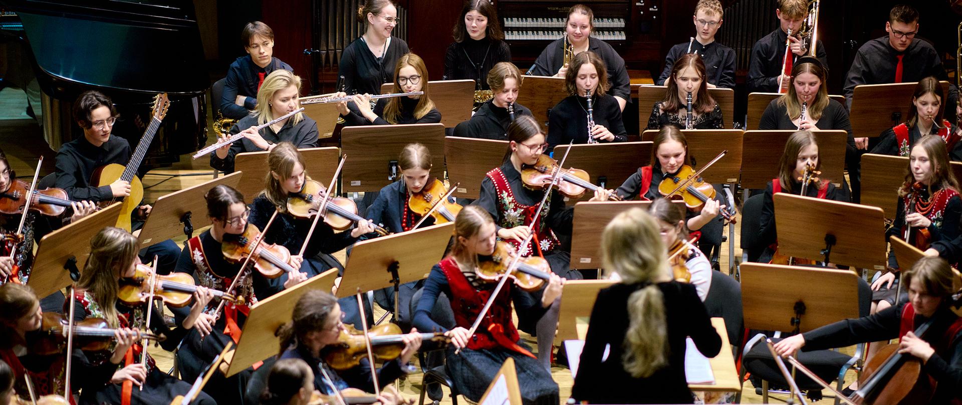 Zdjęcie na sali koncertowej, na pierwszym planie publiczność. Na scenie sali koncertowej orkiestra symfoniczna, przed orkiestrą dyrygentka, Za nimi w tle organy oraz fortepian