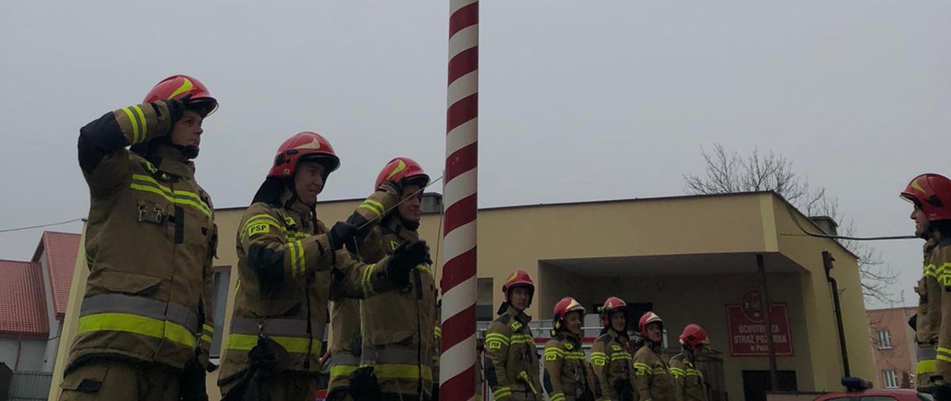Strażacy salutują przy maszcie. Ubrani w ubrania specjalne i czerwone hełmy