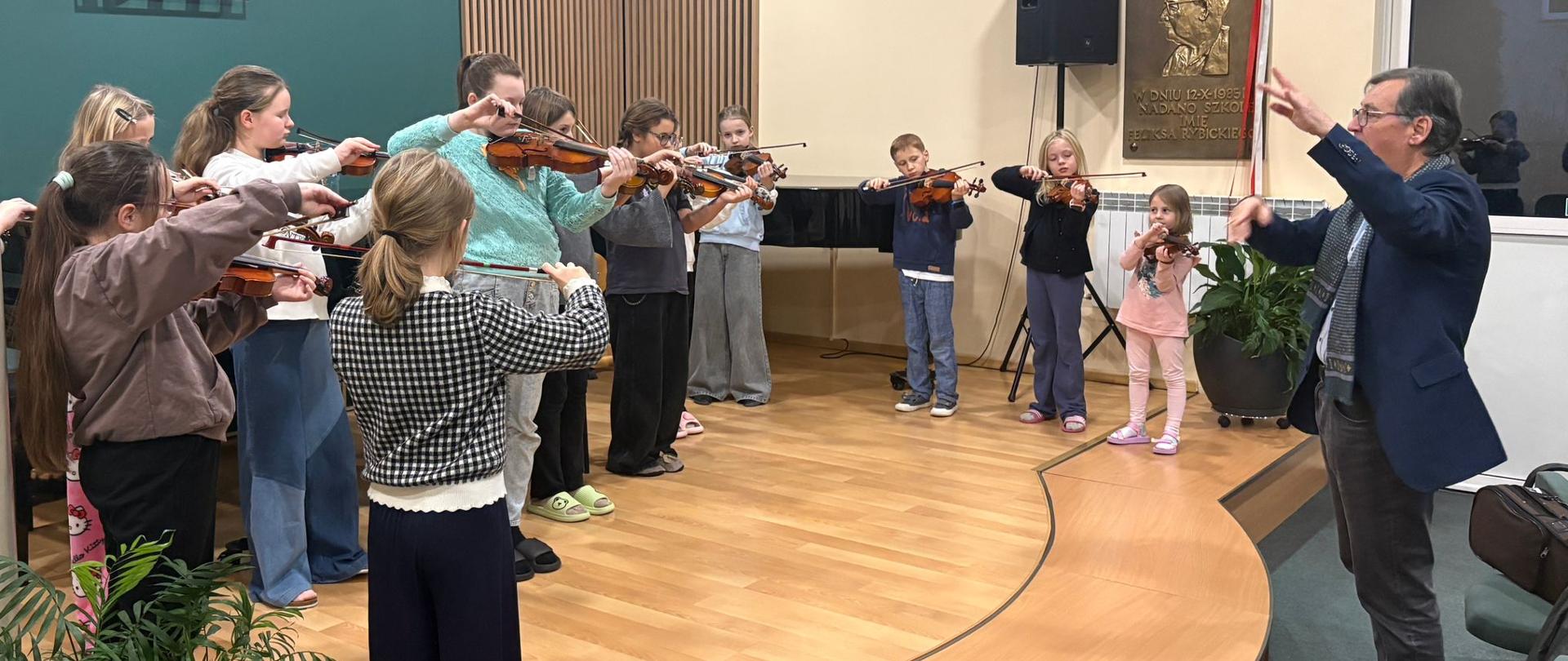 Na zdjęciu widać uczniów na scenie stojących w półokręgu i grających na skrzypcach. Na dole zdjęcia po lewej stronie widać doniczkę z kwiatkami. Pani po prawej stronie na zdjęciu poniżej sceny widać profesora który dyryguje uczniami. W tle widoczna żółta i Zielona ściana.