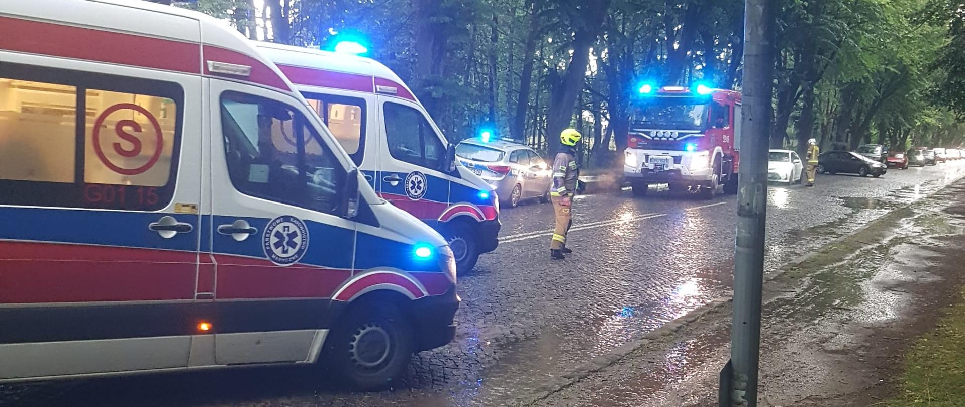 Na zdjęciu widać dwie karetki pogotowia oraz wóz strażacki a przy nim strażaków kierujących ruchem.