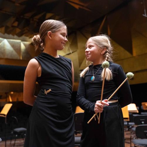 Zdjęcie przedstawia dwie uczennice z dwoma pałkami do gry na instrumencie perkusyjnym we wspólnym zdjęciu portretowym na scenie sali koncertowej filharmonii, inne ujęcie