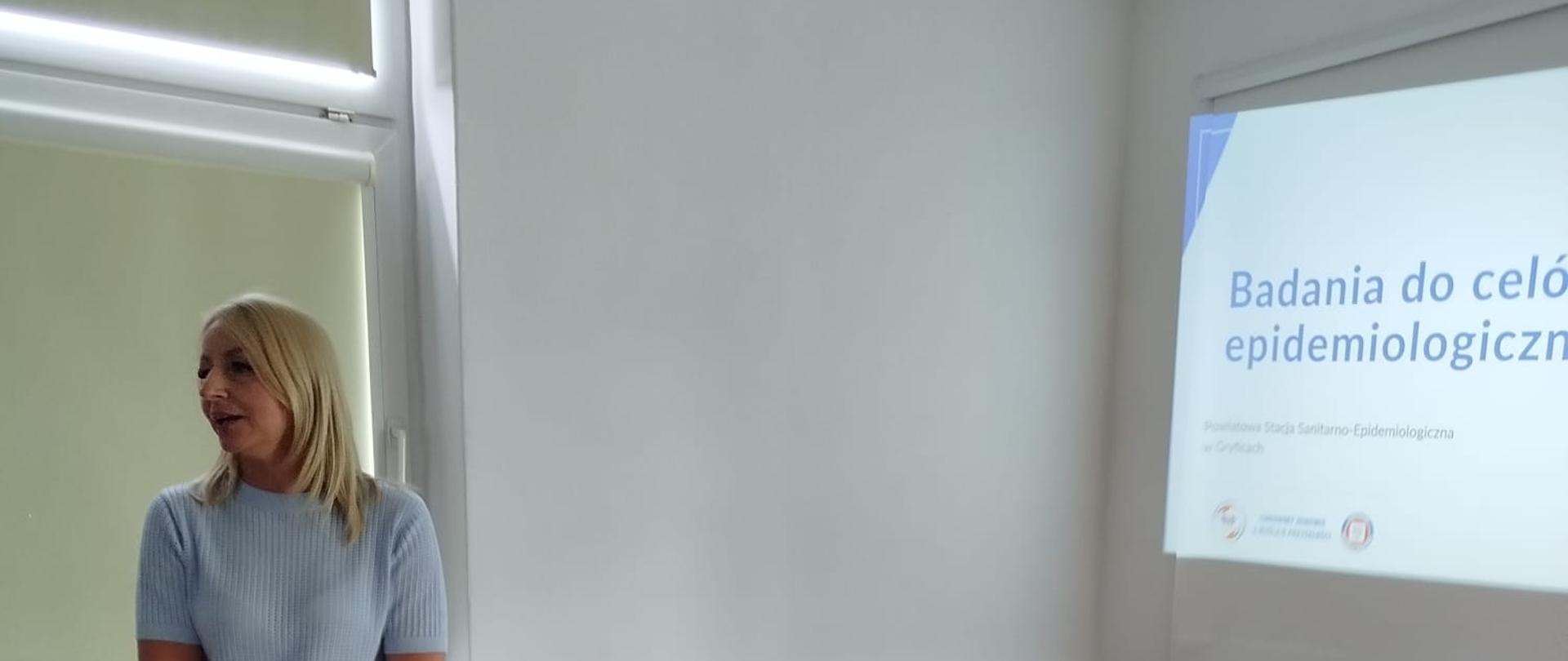 Na zdjęciu sala szkoleniowa z długim stołem konferencyjnym i krzesłami. Przy stole stoi osoba obsługująca laptop i projektor. Na ścianie wyświetlana jest prezentacja zatytułowana „Badania do celów epidemiologicznych”, związana z działalnością Powiatowej Stacji Sanitarno‑Epidemiologicznej w Gryficach. W pomieszczeniu widoczne są okna z opuszczonymi roletami oraz sprzęt multimedialny podłączony do przedłużacza leżącego na stole.