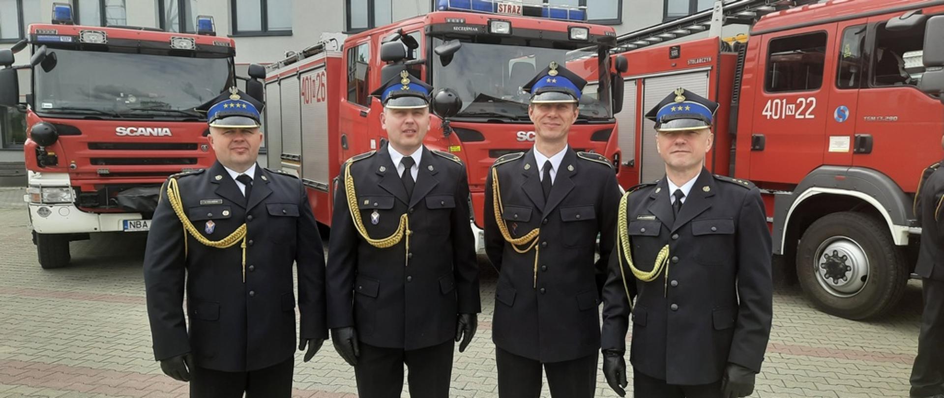 na zdjęciu czterech strażaków w mundurach galowych, w tle, trzy czarowne samochody pożarnicze