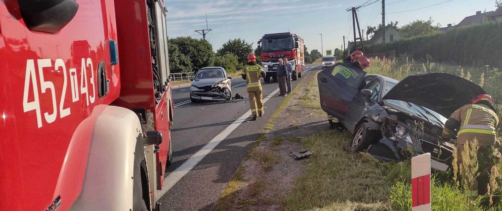 Na jezdni znajduje się samochód gaśniczy, strażak oraz rozbity samochód Peugeot, obok w rowie znajduje się uszkodzony pojazd marki Seat przy którym pracuje 3 strażaków. W oddali na jezdni drugi samochód gaśniczy zaparkowany na jezdni. Pora dzienna.
