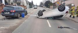 biały samochód leży na dachu na środku drogi z prawej strony na chodniku widać trzech strażaków po drugiej stronie przy krawędzi drogi stoi granatowy samochód przy nim niebieska beczka w oddali droga oraz budynki