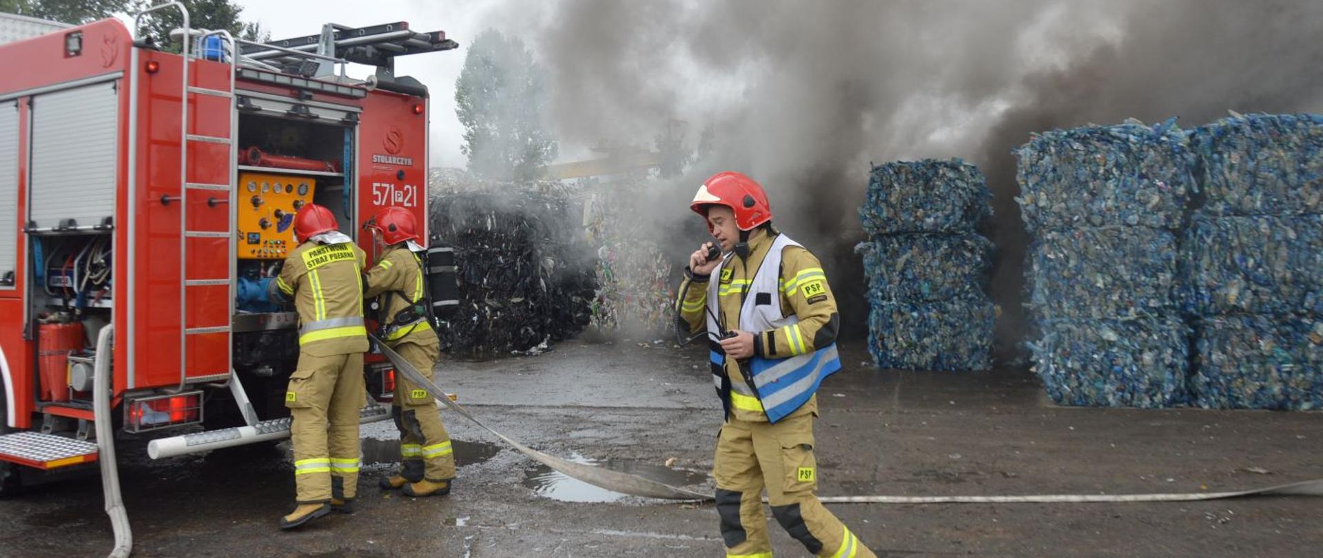 Akcja na składowisku odpadów. Plac, na którym znajdują się pryzmy z odpadami. Na pierwszym planie wóz straży pożarnej oraz strażacy przygotowujący sprzęt do gaszenia pożaru odpadów. Jeden z nich, kierujący działaniem ratowniczym, rozmawia przez radiotelefon. Nad pryzmami unoszą się kłęby dymu. W tle drzewa. 