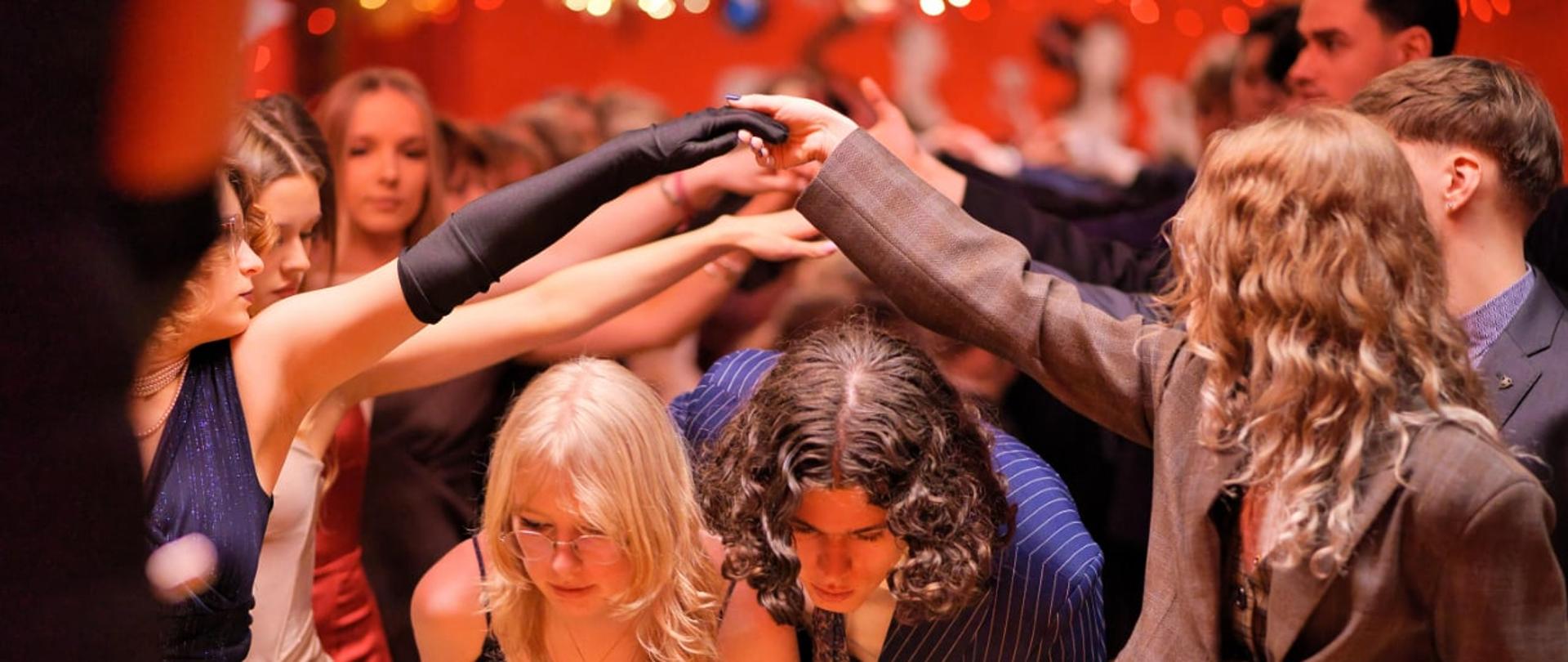 Zdjęcie przedstawia parę młodych ludzi przechodzącą pod „mostem” utworzonym z uniesionych dłoni innych uczestników zabawy, co jest charakterystycznym elementem tradycyjnego tańca – poloneza.
Szczegóły obrazu:
Pierwszy plan: W centrum widać pochyloną parę – dziewczynę w okularach i ciemnej, błyszczącej sukience oraz chłopaka w prążkowanym garniturze. Przechodzą oni pod ramionami innych tancerzy.
Tancerze tworzący szpaler: Po lewej stronie widać dziewczynę w długiej, czarnej rękawiczce, a po prawej osobę w brązowej marynarce. Ich dłonie są złączone wysoko nad głowami przechodzącej pary.
Tło: W oddali widać tłum innych uczestników balu (prawdopodobnie studniówki) oraz czerwone dekoracje ścienne. Oświetlenie jest ciepłe i nastrojowe, w odcieniach pomarańczu i czerwieni.
Ubiór: Wszyscy uczestnicy są ubrani w stroje wieczorowe – garnitury, suknie balowe i eleganckie dodatki.
Fotografia oddaje dynamikę i uroczysty charakter balu maturalnego uczniów lubelskiego „Plastyka”.
