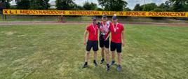Trzech mężczyzn, dwóch w czerwonych koszulkach i jeden w koszulce z biało-czerwonym wzorem, stoi na trawiastym boisku z medalami na szyjach. Za nimi rozciąga się baner z napisem „XLI MIĘDZYNARODOWE MISTRZOSTWA POLSKI W SPORCIE POŻARNICZYM”.