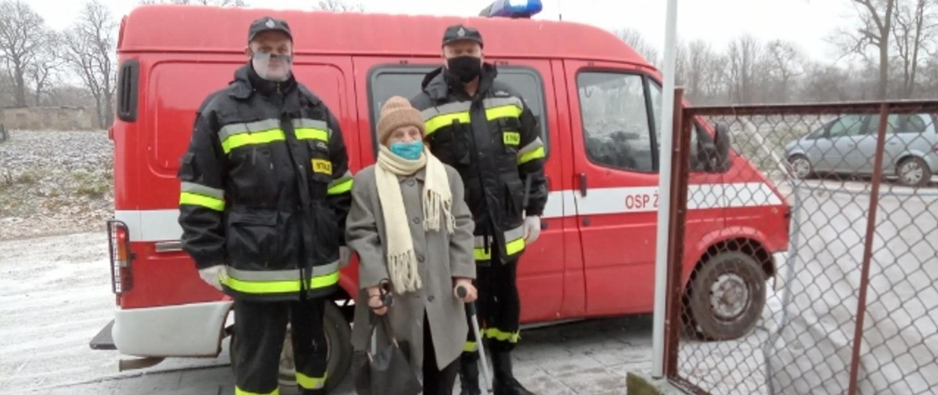 Zdjęcie pamiątkowe. Na tle samochodu pożarniczego stoją dwaj strażacy w towarzystwie stojącej pomiędzy nimi starszej kobiety poruszającej się o kulach. Dowóz na szczepienie.