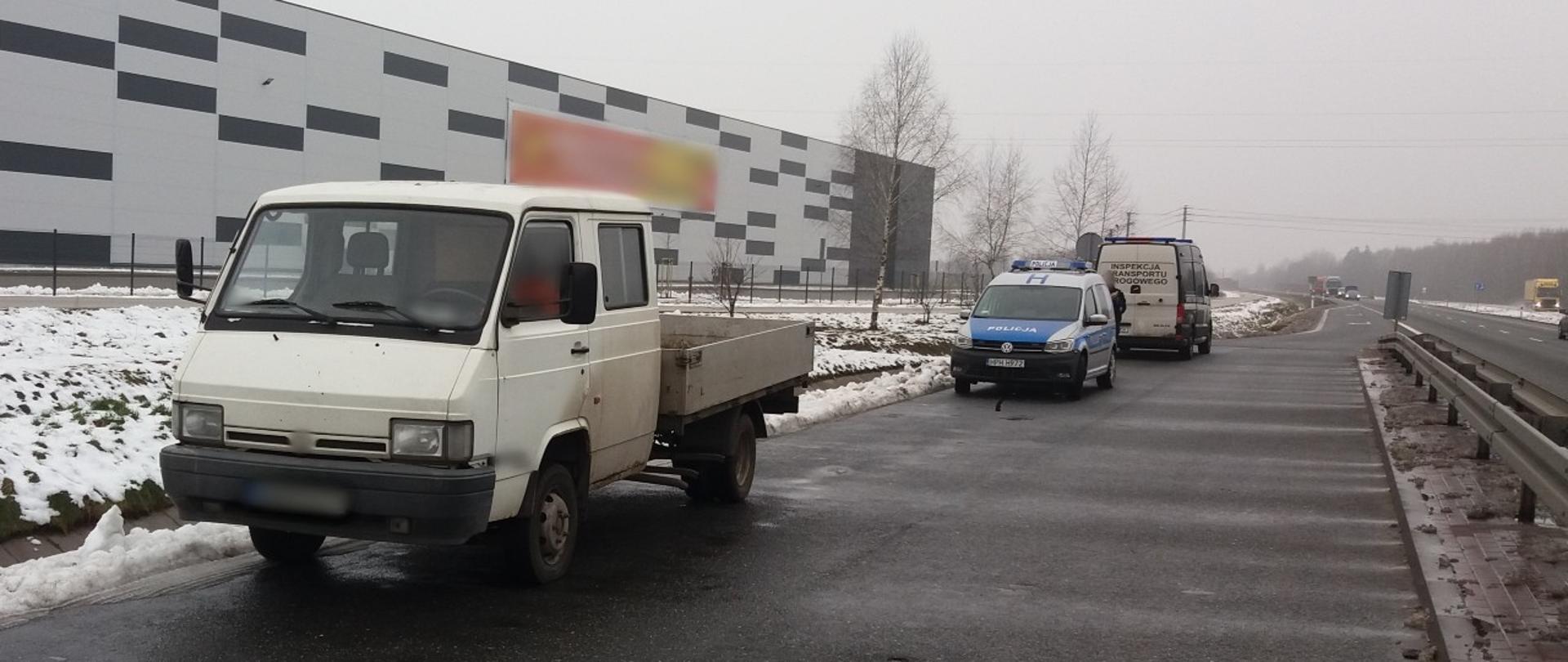 Na pierwszym planie: samochód dostawczy z kabiną załogową i pustą skrzynią ładunkową stoi w zatoce punktu kontrolnego przy krajowej „pięćdziesiątce” . W tle: oznakowany radiowóz Policji i oznakowany furgon mazowieckiej Inspekcji Transportu Drogowego stoją za zatrzymanym do kontroli samochodem dostawczym. 