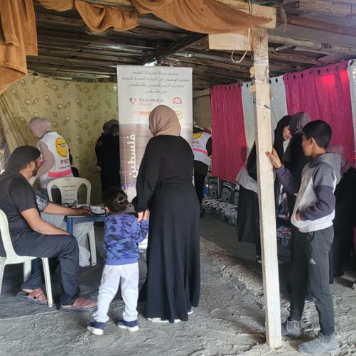 W dużym namiocie kobiety, dzieci i mężczyźni korzystają z pomocy lekarskiej. Widać personel medyczny w kamizelkach z napisem PMM. Mężczyzna ma badane ciśnienie krwi