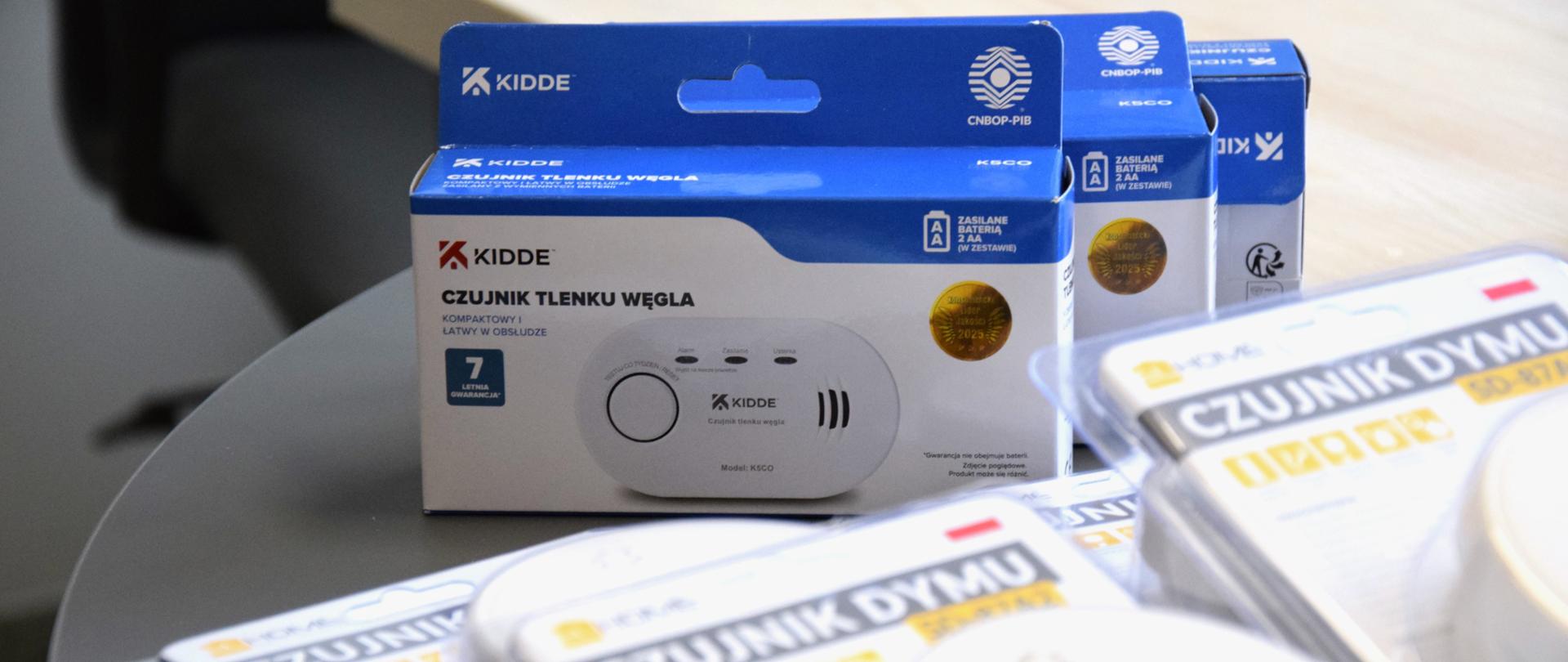 Na fotografii znajdują się czujki tlenku węgla oraz czujki dymu. Wszystkie czujki leżą na szarym, okrągłym stoliku. Wszystkie czujki są w oryginalnych opakowaniach. Czujki obejmują większą powierzchnię zdjęcia, jest wykonane na nie zbliżenie. Czujki dymu leżą przed czujkami tlenku węgla i są rozmyte. Czujki tlenku węgla w biało niebieskim opakowaniu są bardzo wyraźne.