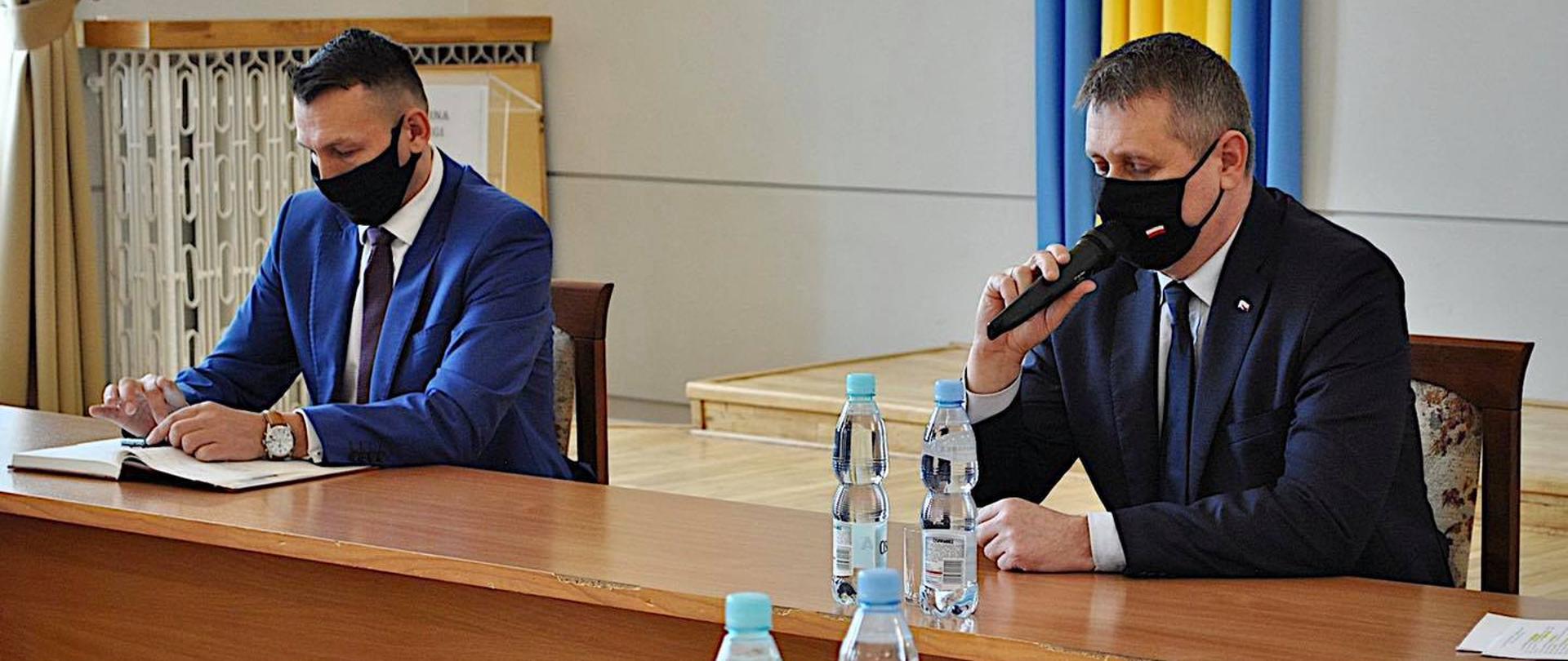 Dwóch eleganckich mężczyzn siedzących przy brązowym stole. Wicewojewoda Standowicz przemawia do mikrofonu. Przed nim dwie butelki wody. 