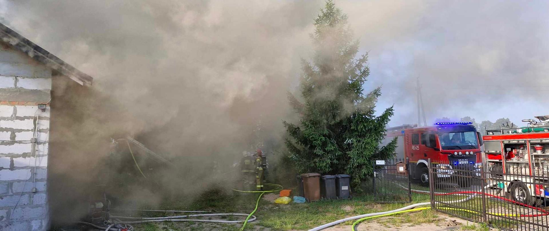 Widoczny gęsty dym wydobywający się nad budynkiem mieszkalnym. Na placu manewrowym przed obiektem duża ilość węży pożarniczych. Strażacy prowadzą działania gaśnicze.