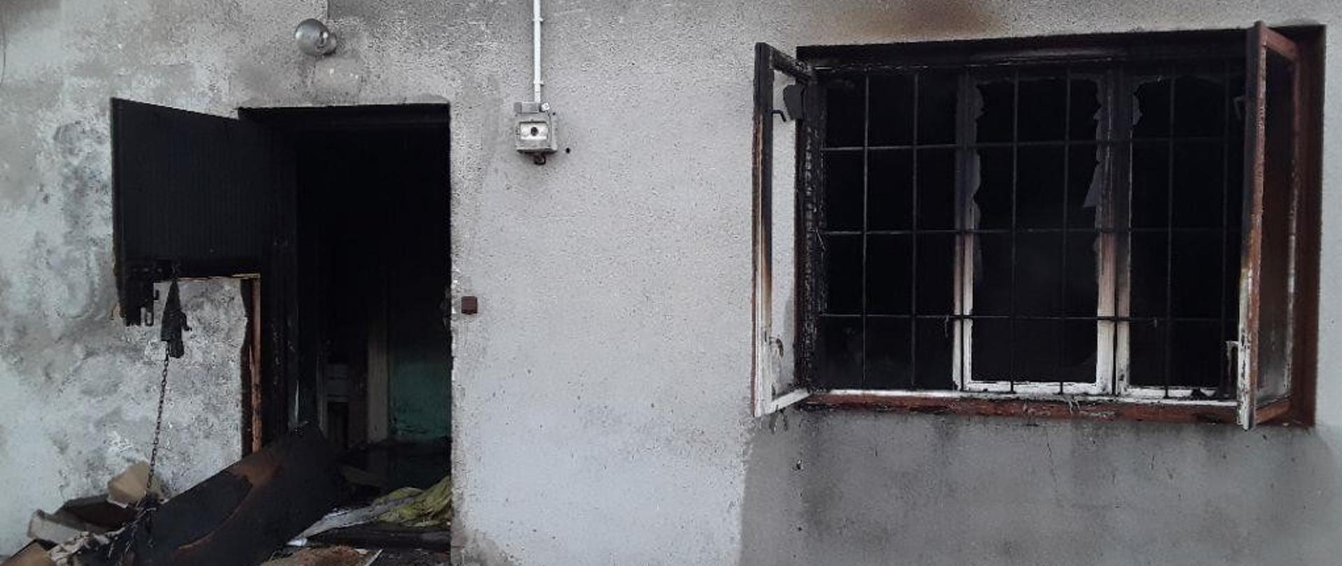 Drzwi wejściowe oraz okno budynku, w którym doszło do pożaru. W oknie budynku widoczne kraty. Drzwi zniszczone. Stolarka okienna zniszczona przez ogień.