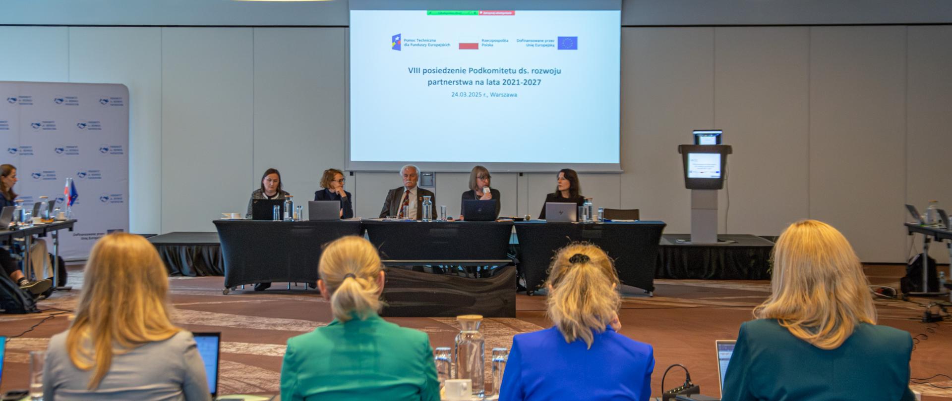 Grupa osób siedzi przy stole. W oddali na ścianie widać ekran. 