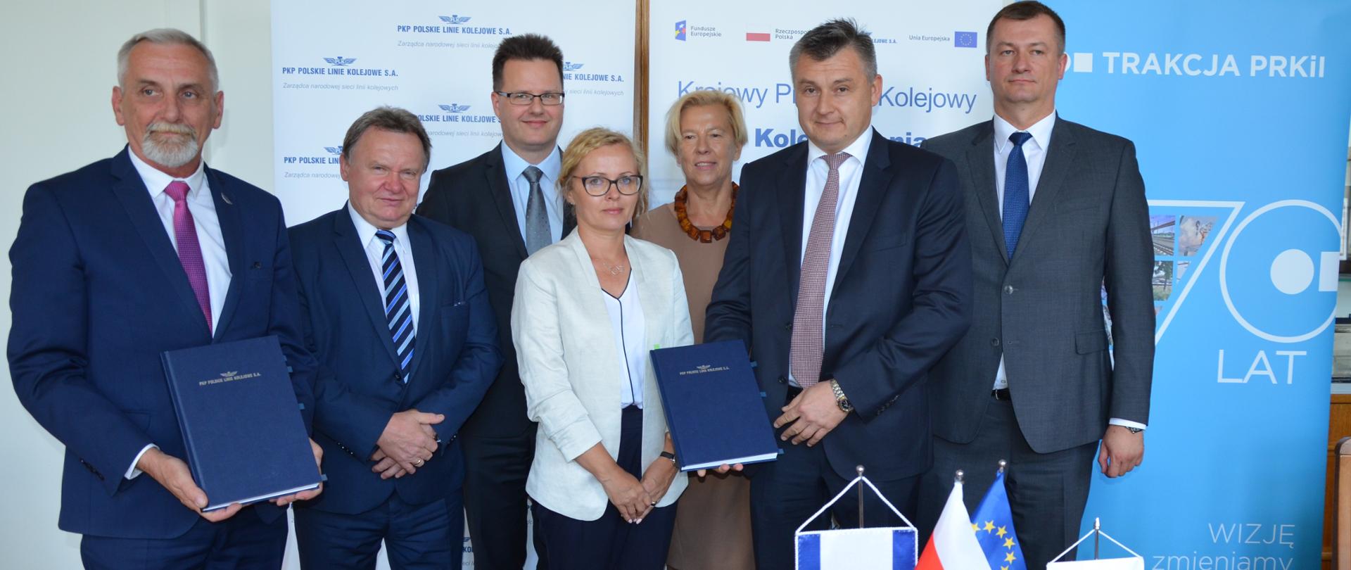 Na zdjęciu stoi siedem osób, dwie z nich trzymają w ręku umowy 