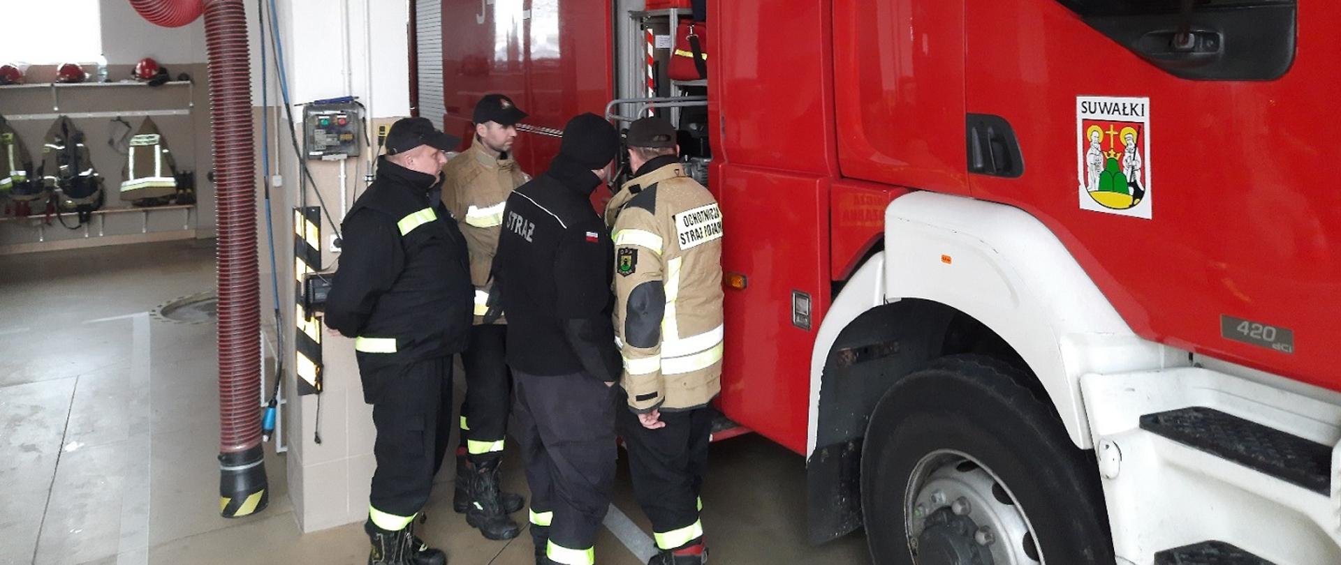 Zdjęcie obrazuje strażaków w garażu oglądających sprzęt pożarniczy znajdujący się na samochodzie gaśniczym 