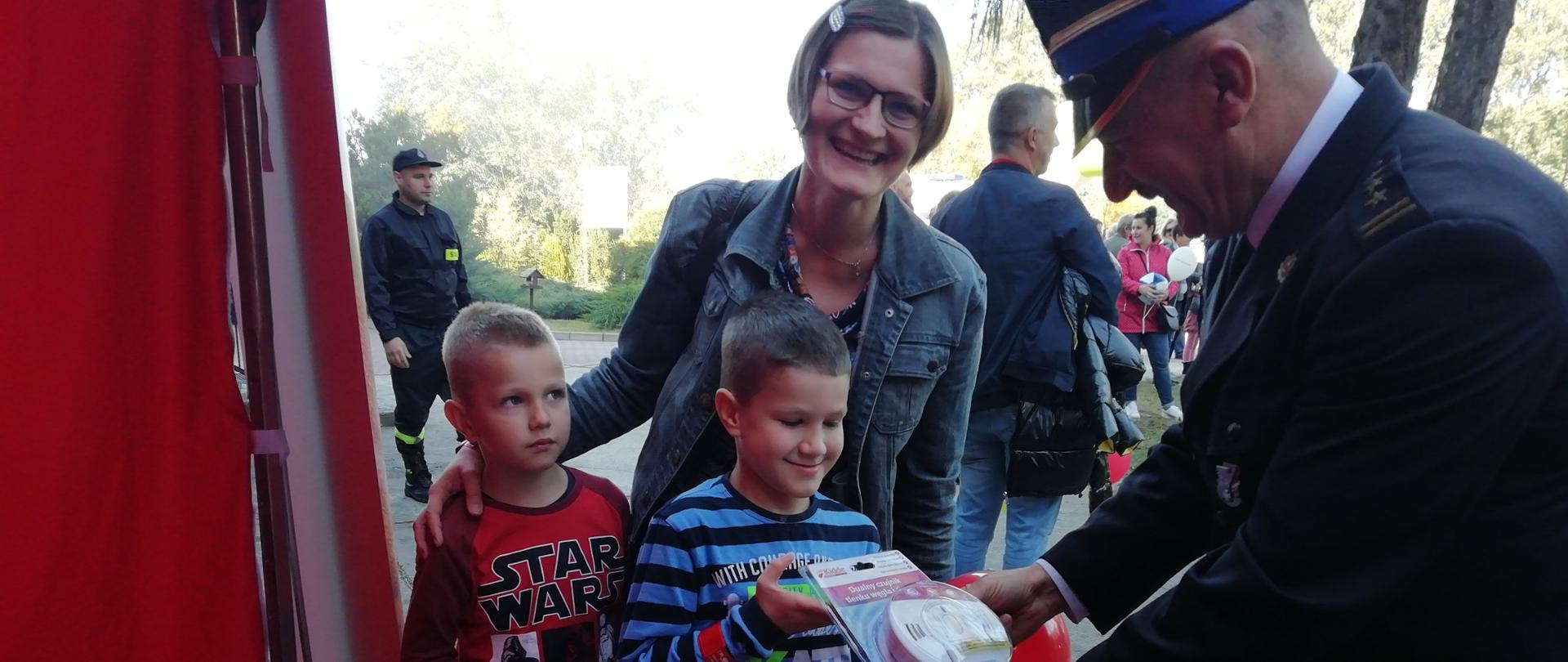 Strażak wręcza chłopcu czujnik czadu. Za chłopcem stoi kobieta, która obejmuje drugiego chłopca. W tle widać strażaka i mężczyznę stojącego tyłem. Z lewej strony widać balony i lizaki. 