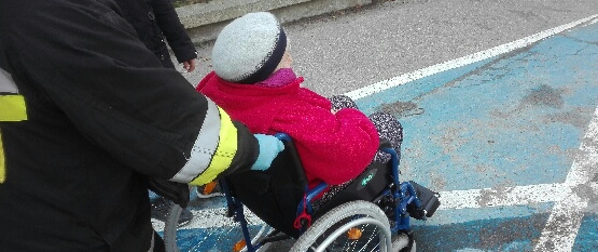 Obraz przedstawia strażaka prowadzącego starszą osobę poruszającą się na wózku inwalidzkim na szczepienie przeciwko koronawirusowi.