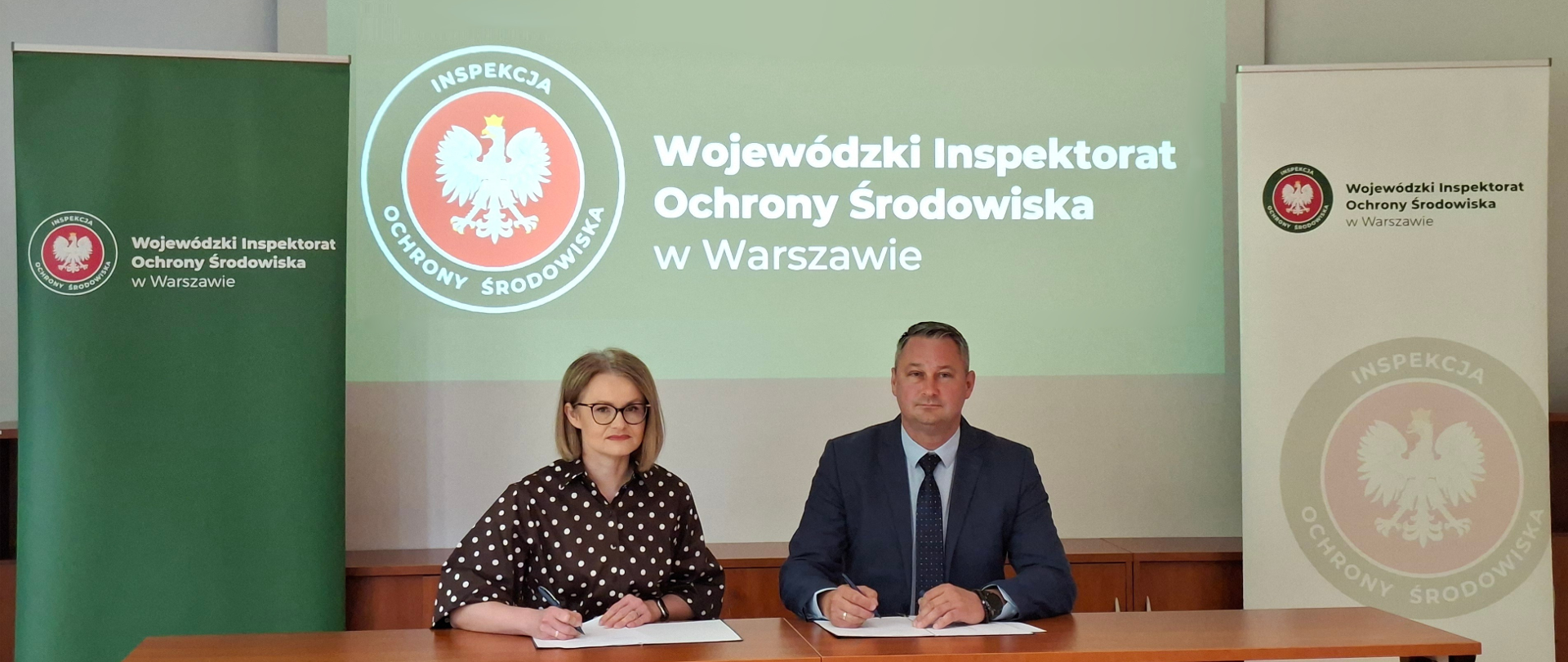 Przy stole na sali konferencyjnej siedzą Mazowiecki Wojewódzki Inspektor Ochrony Środowiska Krzysztof Cząstkiewicz i Mazowiecki Wojewódzki Lekarz Weterynarii Izabela Floryszczyk. Przedstawiciele obu inspekcji mają długopisy w dłoniach i są gotowi do podpisania leżących przed nimi kopii porozumienia. Patrzą się w obiektyw aparatu.