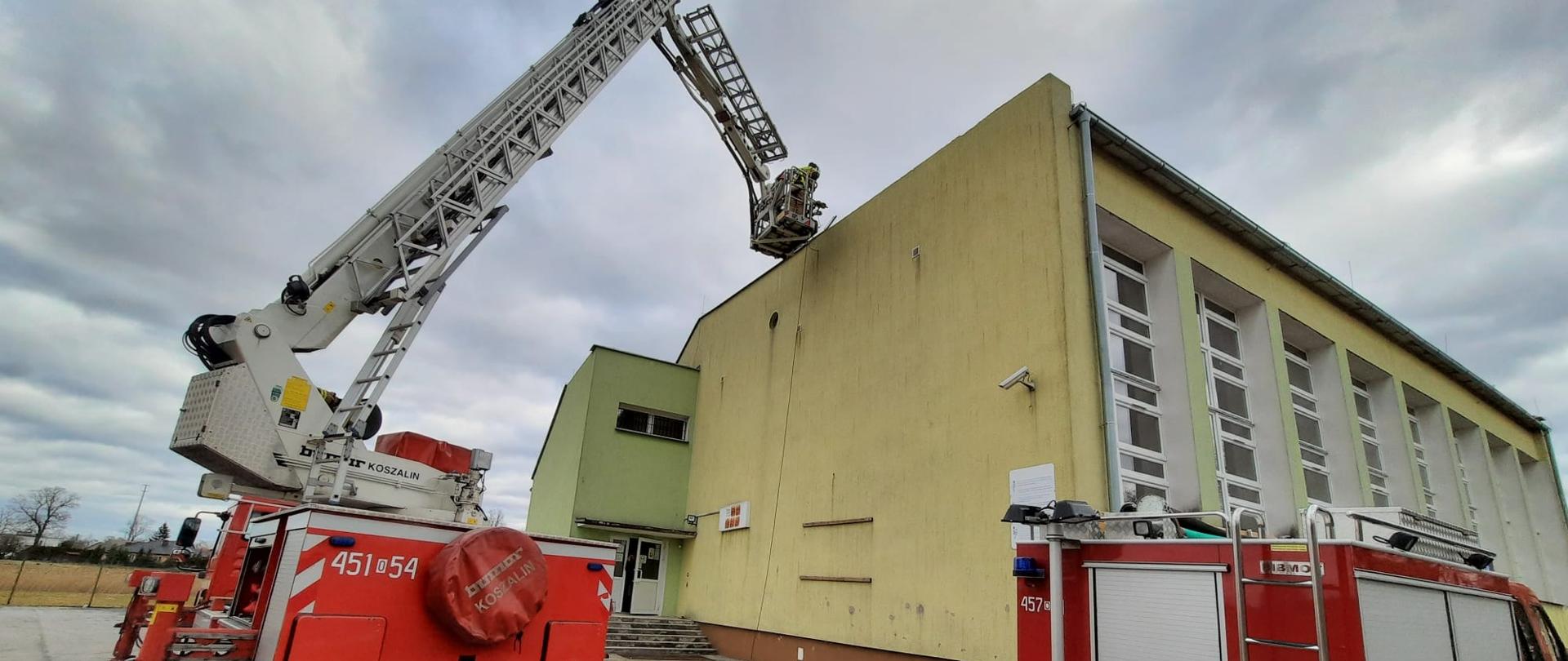 Strażacy PSP i OSP usuwają skutki silnego wiatru, który przeszedł nad powiatem namysłowskim. W lewej części widoczny podnośnik hydrauliczny JRG Namysłów w trakcie działań przy budynku szkoły. Kosz ratowniczy pojazdu usadowiony na wysokości dachu obiektu sali gimnastycznej. Po prawej widoczny samochód pożarniczy koloru czerwonego.