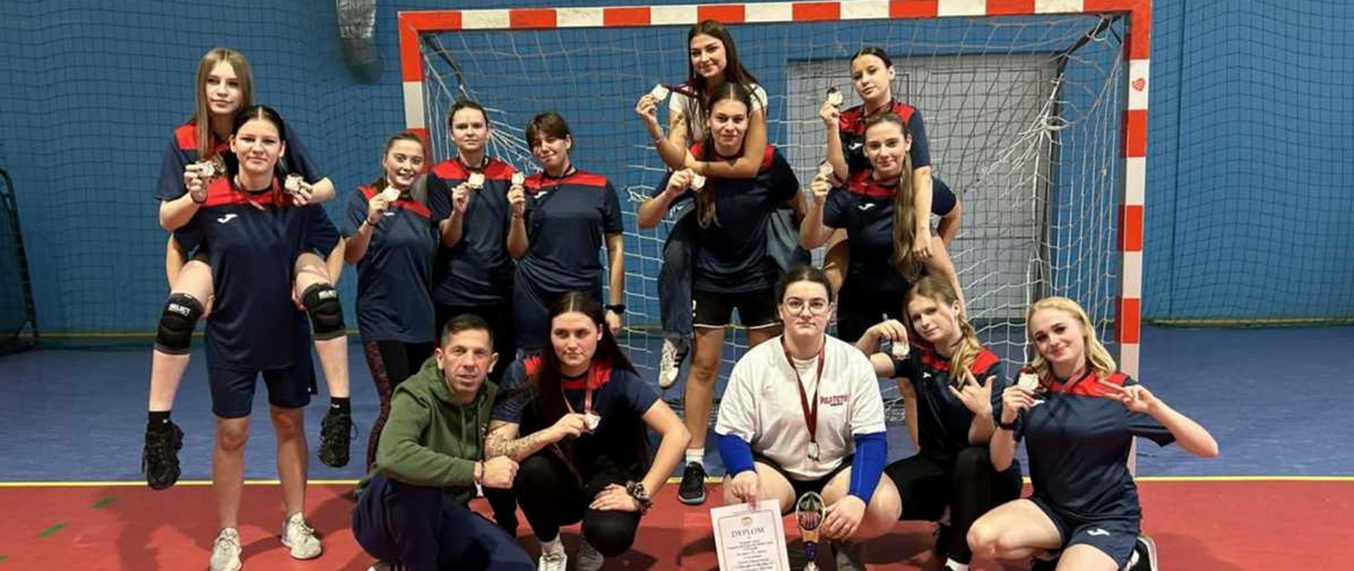 Zdjęcie przedstawia uśmiechniętą drużynę sportową, składającą się z kilkunastu młodych kobiet oraz trenera, pozujących do wspólnego zdjęcia w hali sportowej, na tle bramki.
Wygląd i ubiór
Drużyna ubrana jest w jednolite, ciemnoniebieskie stroje sportowe (koszulki i spodenki) z czerwonymi akcentami. Wszystkie zawodniczki trzymają w dłoniach medale zawieszone na szyi lub uniesione w górę.
Trener (mężczyzna) stoi po lewej stronie, ubrany w szary sweter z kapturem (hoodie) i ciemne spodnie.
Zawodniczka siedząca na środku pierwszego rzędu, w białej koszulce, trzyma w rękach dyplom w ramce oraz mały puchar stojący obok niej na podłodze, co sugeruje zwycięstwo w zawodach.
Pozycje i tło
Grupa jest ułożona w dwóch rzędach: stojącym z tyłu i klęczącym z przodu. Niektóre dziewczyny stoją na plecach koleżanek (na barana).
Tło stanowi boisko sportowe z czerwoną nawierzchnią i białymi liniami. Bezpośrednio za drużyną znajduje się metalowa bramka sportowa z siatką. W głębi widać ściany hali w kolorach niebieskim i żółtym, oraz fragment podłogi w kolorze jasnego drewna.
