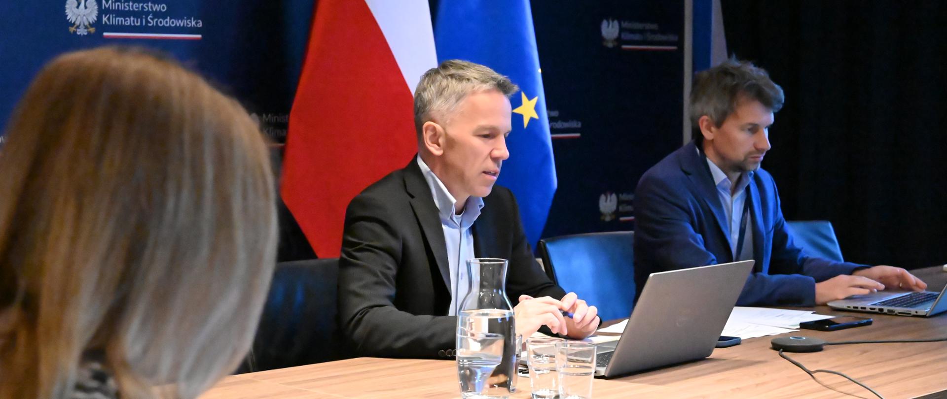 Trzy osoby siedzą z komputerami przy biurku. W centrum na tle flagi polskiej i EU siedzą członkowie Międzyresortowego Zespołu ds. Odry