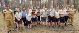 Wesoła grupa harcerzy (w krótkich granatowych spodenkach, w koszulach oliwkowych i zielono żółty chustach zawiązanych na szyi) wszyscy stoją w lesie sosnowym, po lewej i prawej strażacy z Komendy Powiatowej Państwowej Straży Pożarnej w Białobrzegach w piaskowych mundurach.