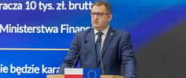 Mężczyzna przemawia z mównicy. W tle prezentacja o KSeF.