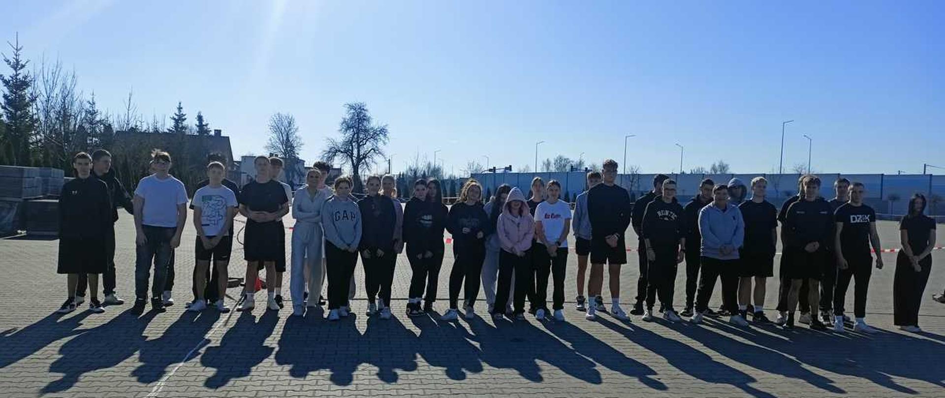 Na zdjęciu widać grupę około trzydziestu sześciu uczniów stojących na parkingu szkolnym przy hali sportowej. Uczestnicy ustawieni są w jednym rzędzie, obok siebie tuż przed startem lub po zakończeniu biegu. Wszyscy mają na sobie strój sportowy – bluzy, dresy, legginsy, koszulki oraz sportowe obuwie.
