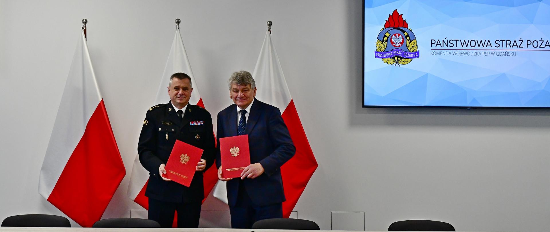 Pomorski komendant wojewódzki Państwowej Straży Pożarnej oraz wójt gminy Kobylnica trzymają czerwone teczki. Za nimi stoją flagi państwowe oraz wyświetlane jest logo Państwowej Straży Pożarnej.