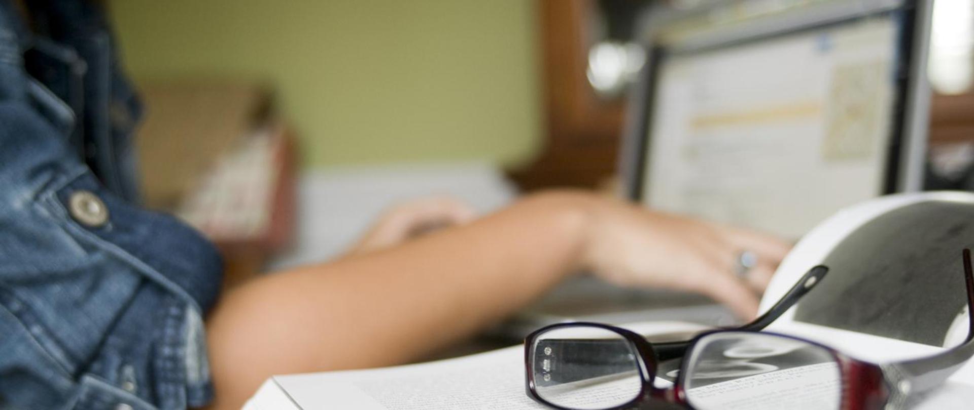 Zdjęcie przedstawia osobę pracującą przy laptopie z otwartą książką i okularami na pierwszym planie.