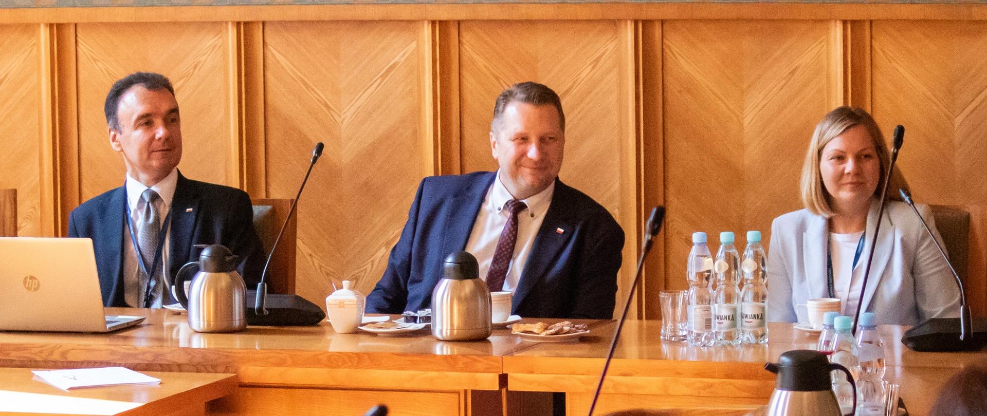 Minister Czarnek siedzi za drewnianym stołem, obok niego siedzi ubrana w jasne ubranie kobieta i mężczyzna w garniturze.