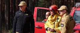 Czterech mężczyzn w mundurach służb ratowniczych stoi obok czerwonego pojazdu terenowego na tle lasu. Jeden z nich ubrany w żółty strój strażaka i kask, pozostali w kurtkach z emblematami Państwowej Straży Pożarnej i Lasów Państwowych. Trwają rozmowy lub ustalenia, prawdopodobnie podczas działań operacyjnych w terenie leśnym.