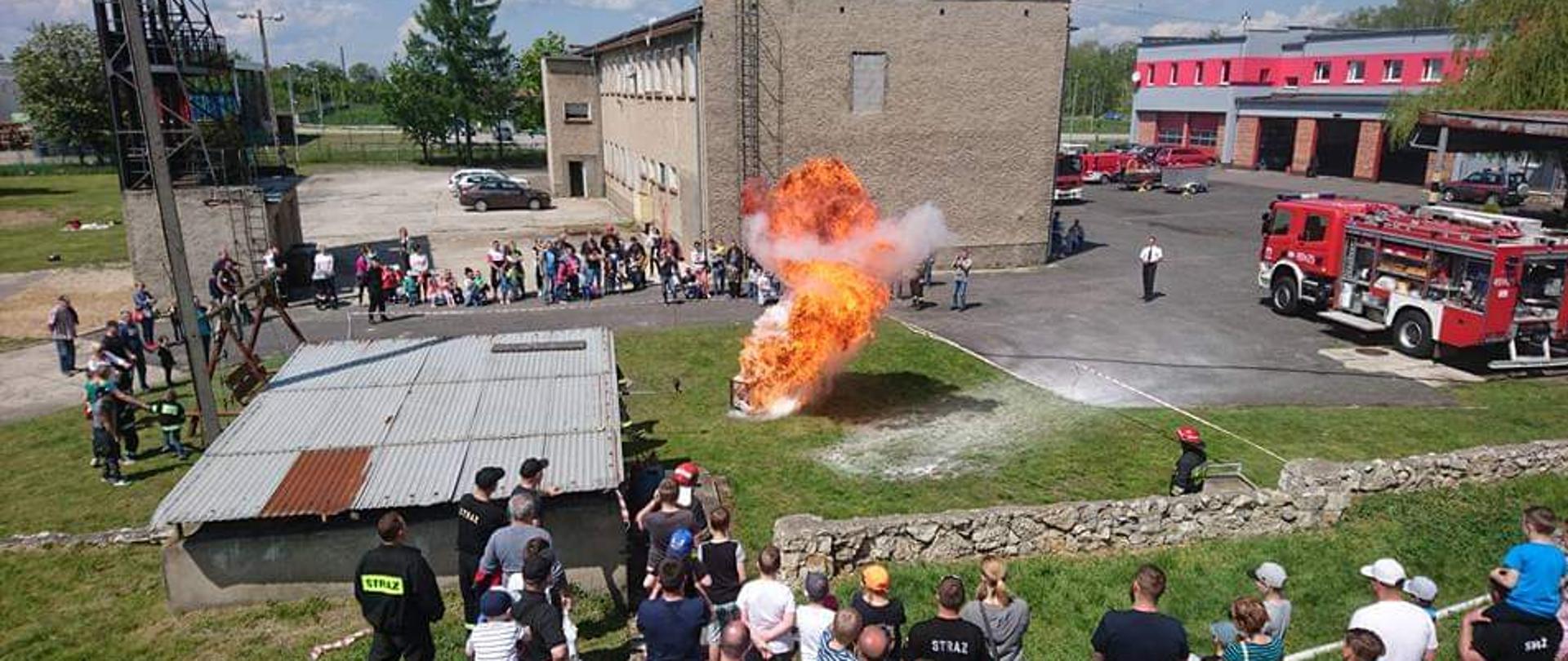 Zdjęcie przedstawia kłęby dymu i ognia oraz osoby zgromadzone wokół miejsca palącego się oleju na tle wspinalni, budynku komendy oraz pojazdów pożarniczych