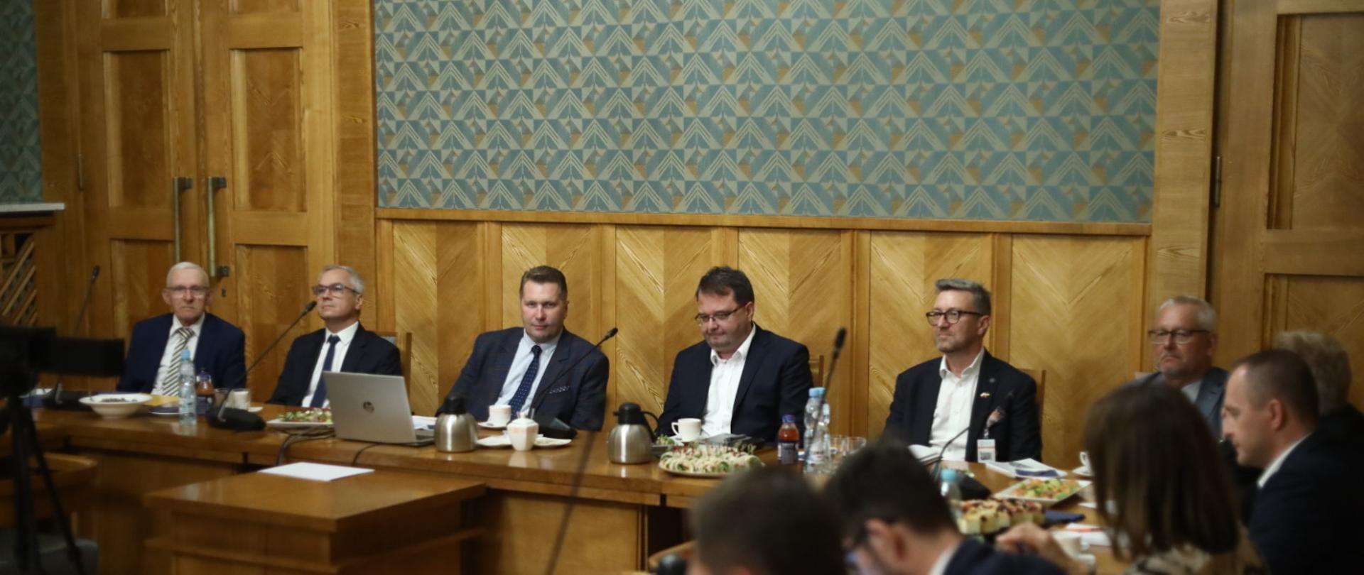 Dwunastu mężczyzn siedzi przy stole, wszyscy słuchają. Na stole dzbanki, filiżanki, woda, ciastka i owoce