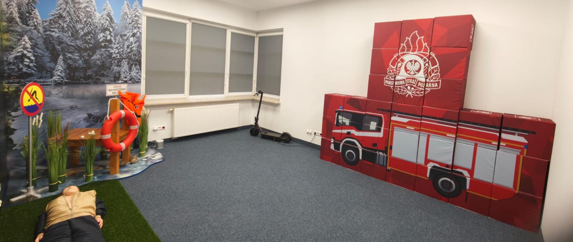 Sala edukacyjna poświęcona bezpieczeństwu i pracy strażaków. Na ścianie stoją czerwone moduły z wizerunkiem wozu strażackiego i emblematem Państwowej Straży Pożarnej. Po lewej stronie znajduje się aranżacja zimowego lasu z fantomem osoby leżącej na ziemi, kołem ratunkowym i oznakowaniem ostrzegawczym.