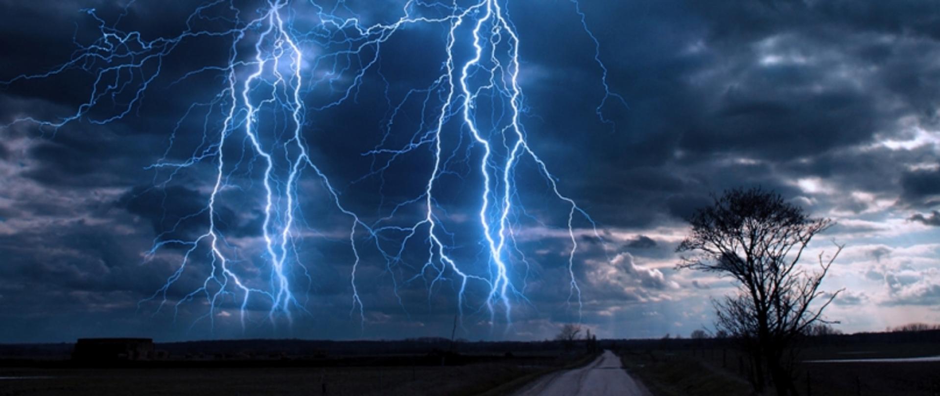 Pioruny uderzające z ciemnego nieba nad pustą drogą i polami, z samotnym drzewem po prawej stronie.