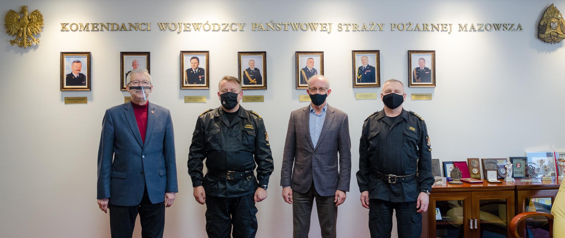 Zdjęcie przedstawia cztery osoby, w tym dwóch strażaków w umundurowaniu dowódczo-sztabowym. W tle ściana z portretami byłych komendantów wojewódzkich PSP Mazowsza.