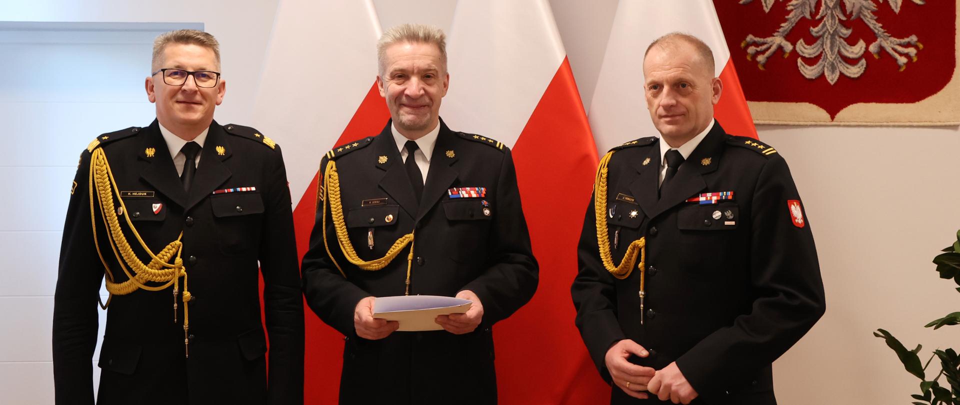 Zdjęcie pamiątkowe. Trzech mężczyzn w mundurach pozujący na tle flag narodowych i godła