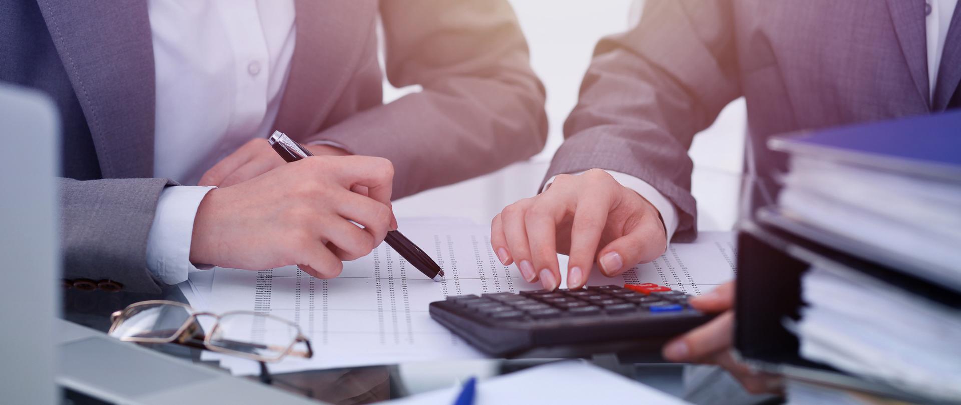 Dwie osoby siedzą przy biurku, obok kalkulator, długopisy, komputer, segragatory
