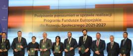 Zdjęcie zbiorowe, duża grupa elegancko ubranych ludzi trzymających ciemnozielone teczki z godłem stoi w rzędzie pod ścianą z napisem Podpisanie porozumień w sprawie realizacji Programu Fundusze Europejskie dla Rozwoju Społecznego 2012-2027.