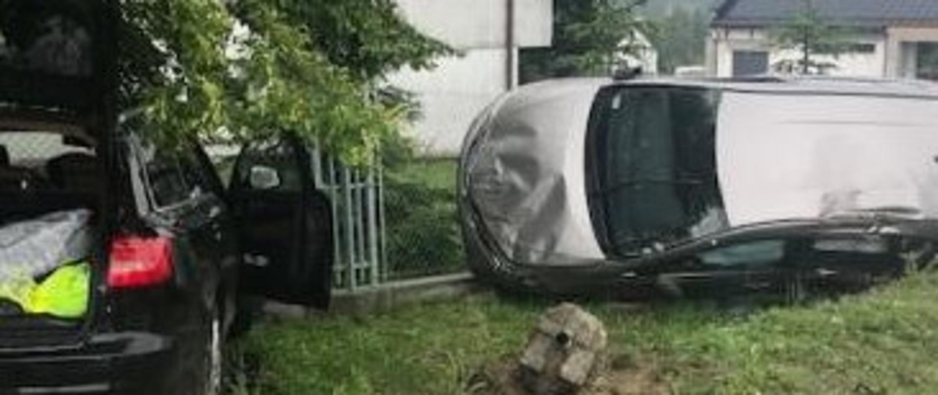 wypadek dwóch samochodów osobowych. Jedno auto widoczny tył, drugie auto znajduje się na boku z widocznymi uszkodzeniami. Auta znajdują się poza drogą.