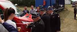 Zdjęcie przedstawia przedstawicieli władz państwowych oraz władz Państwowej i Ochotniczej Straży Pożarnej podczas uroczystego przecięcia biało-czerwonej wstęgi, przy przekazaniu nowego samochodu GLBM 02/05/20 Ford Transit dla Ochotniczej Straży Pożarnej w Świeżawach. 
