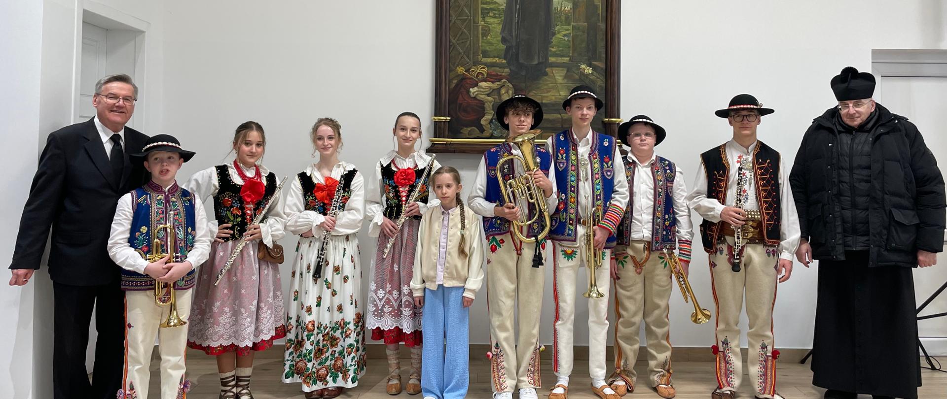 Zdjęcie przedstawia dwójkę młodych muzyków podczas wspólnego występu w sali koncertowej: Wykonawcy: Po lewej stronie widać dziewczynkę grającą na skrzypcach, a po prawej chłopca grającego na trąbce. Oboje są ubrani w stroje galowe i wykazują duże skupienie na swoich instrumentach. Instrumenty: Skrzypaczka trzyma instrument w pozycji do gry, a trębacz trzyma złocistą trąbkę. Przed muzykami ustawione są profesjonalne pulpity z nutami. Tło: W tle widoczny jest imponujący prospekt organowy z pionowymi, metalowymi piszczałkami, co podkreśla podniosłą atmosferę wydarzenia w szkole muzycznej. To dynamiczne ujęcie dokumentuje współpracę młodych artystów na scenie podczas wspólnego muzykowania.